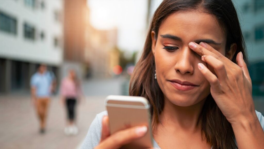 Mujer joven con molestias oculares mientras usa el celular, posible síntoma de visión borrosa frecuente en zonas urbanas de Santiago del Estero (Argentina).
