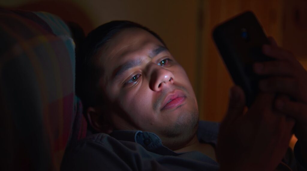 Hombre con fatiga visual mirando su celular en la oscuridad, posible causa del temblor involuntario en el ojo por sobreexposición a pantallas.